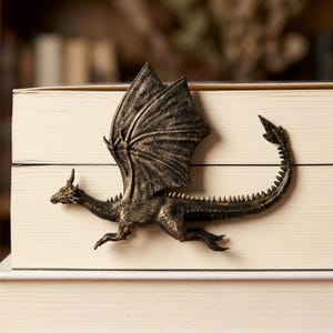 May include: A bronze-coloured dragon bookmark with detailed wings, a long spiked tail, and textured scales. The dragon is positioned horizontally across the spine of a closed book, featuring a metallic finish with dark highlights in the crevices.