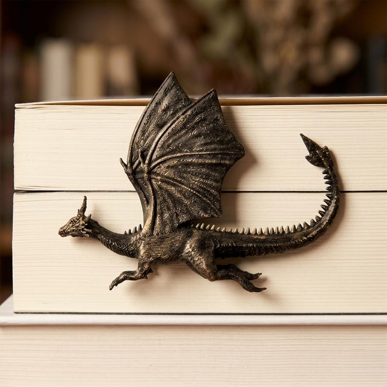 May include: A bronze-coloured dragon bookmark with detailed wings, a long spiked tail, and textured scales. The dragon is positioned horizontally across the spine of a closed book, featuring a metallic finish with dark highlights in the crevices.