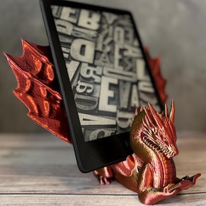 Peut inclure: Un support de tablette en forme de dragon rouge et or. Le dragon a des écailles et des ailes détaillées, tenant une tablette noire affichant une page de texte. Le support est conçu pour maintenir la tablette à la verticale.