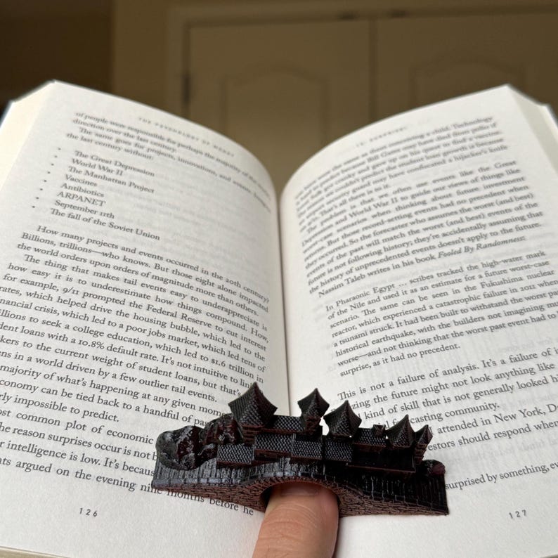 May include: A dark brown 3D-printed book page holder shaped like a Japanese pagoda. The holder is placed on a book, with the pages open to text. The holder is designed to slip over a finger.