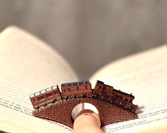 May include: A brown train book page holder with a bridge design. The train is made of three connected cars and a locomotive. The book page holder is designed to fit over a finger.