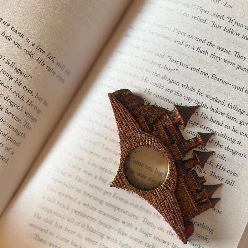 May include: A detailed, bronze-colored metal bookmark shaped like a castle, resting on an open book. The bookmark features a circular magnifying glass and the text "Come on, big boy, way too". The book's pages display printed text.