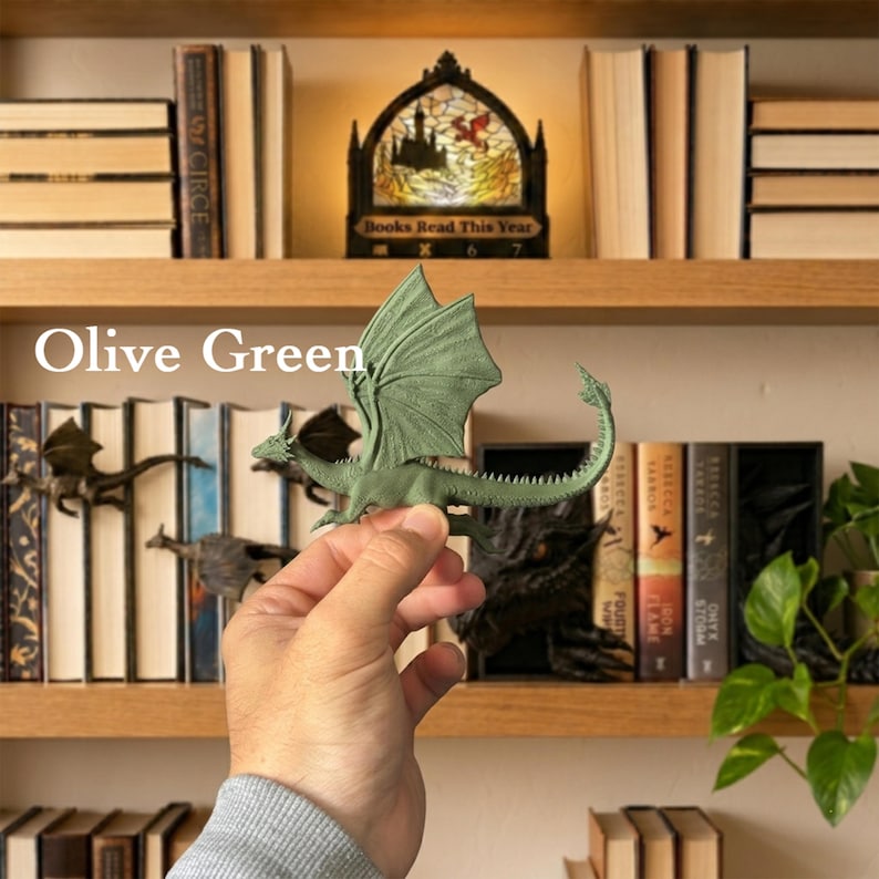 May include: An olive green dragon figurine with detailed wings and a long, curved tail. The dragon is sculpted with textured scales and is held in a position that displays its full profile. The text "Olive Green" is visible on the left side.