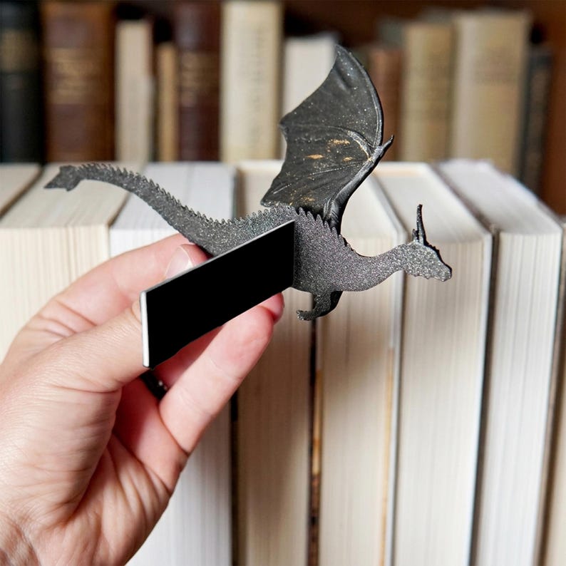 May include: A black dragon-shaped bookmark featuring a textured, glittery surface and a long, pointed tail. The bookmark includes a rectangular black magnetic strip attached to the centre of the dragon's body, designed to clip onto book pages.