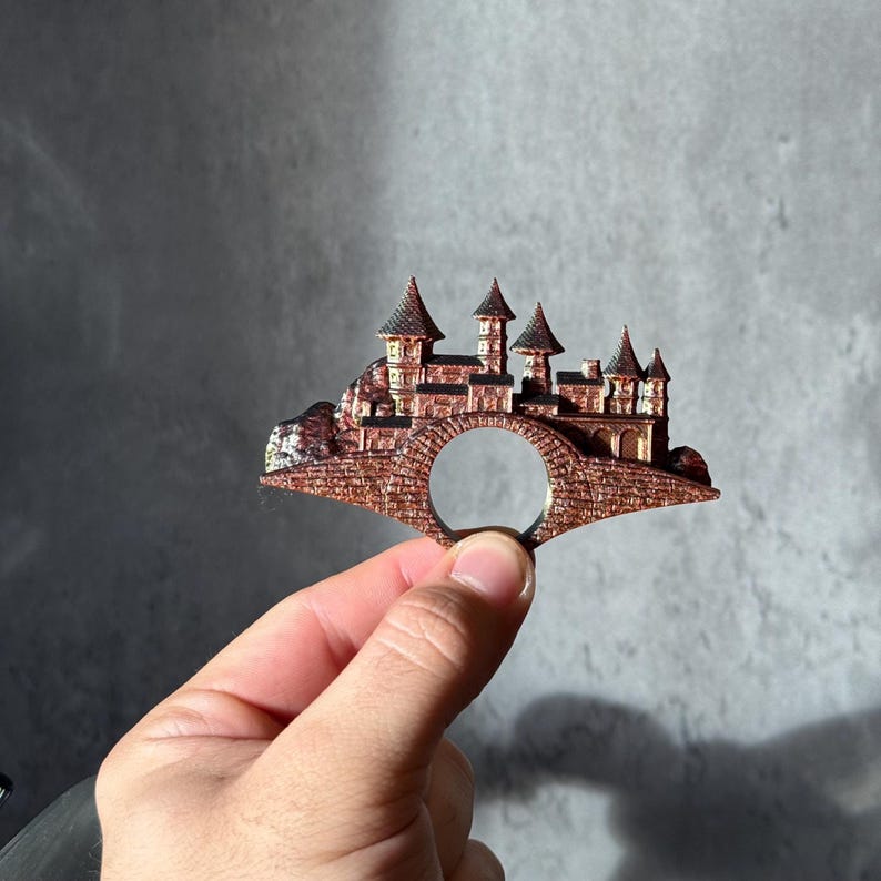 May include: A copper-colored ring featuring a detailed miniature castle design. The castle has multiple turrets and is connected to a bridge with a circular opening. The ring is held against a gray background.