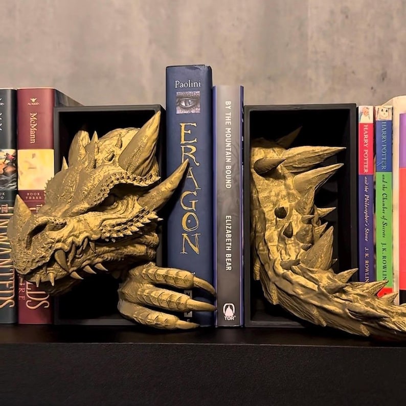 May include: Gold dragon head bookends supporting a row of books. The bookends feature intricate scale and claw details. Books include titles such as "Eragon" and "Harry Potter". The bookends are on a black shelf.