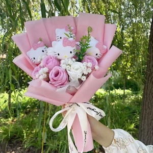 以下が含まれることがあります： ピンクのバラ、白い花、ピンクのリボンが付いた白いハローキティのぬいぐるみで作られた花束。 花束はピンクの紙で包まれ、「Just For You」と書かれた白いリボンで結ばれています。