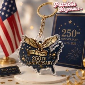 May include: A keychain featuring a gold eagle over a map of the United States, commemorating the 250th anniversary from 1776-2026. The keychain has a gold chain and ring. The background includes an American flag and a navy blue card with the text "250th Anniversary".