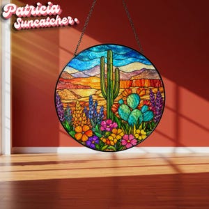 May include: A circular stained glass suncatcher featuring a desert landscape with a tall green saguaro cactus, a prickly pear cactus, and colourful wildflowers in shades of orange, pink, purple, and yellow against a blue sky and orange hills.