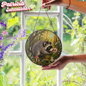 May include: A circular suncatcher featuring a detailed illustration of a raccoon in a forest setting with mushrooms and flowers. The artwork is framed in a thin black metal border and hangs from a delicate metal chain. The text Patricia Suncatcher is visible in the upper left corner.