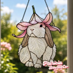 May include: Stained glass suncatcher depicting a white rabbit with a pink flower hat, suspended by a chain. The rabbit has long ears. The text "Patricia BourkeCo" is at the bottom.