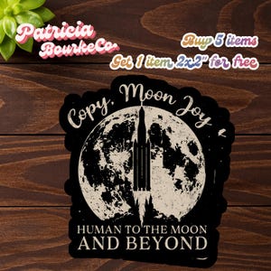 Pode incluir: Adesivo preto com uma ilustração branca de um foguete lançando da lua. O texto no adesivo diz "Copy. Moon Joy. HUMAN TO THE MOON AND BEYOND." O adesivo tem uma borda preta e está em uma superfície de madeira. O texto "Patricia Bourke Co."
