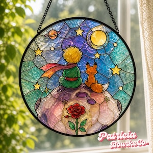 Op de afbeelding: Rond gebrandschilderd glazen raamkunst met een scène uit De Kleine Prins. Het kunstwerk toont de Kleine Prins, een vos, een roos en een nachtelijke hemel met sterren, een maan en planeten. Het heeft een zwart metalen frame en een ophangketting.