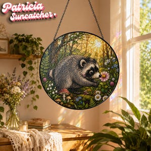 May include: A circular suncatcher featuring a detailed illustration of a raccoon in a forest setting with mushrooms and flowers. The artwork is enclosed in a black metal frame and suspended by a black chain. The text Patricia Suncatcher is visible.