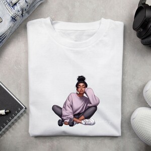 May include: White t-shirt featuring a seated illustration of a person in a lavender sweatshirt and gray leggings, holding a dumbbell. The person has dark hair in a bun and is wearing white sneakers. The illustration has a modern, artistic style.