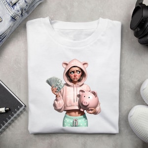 May include: White t-shirt featuring a cartoon illustration of a person in a pink hoodie with bear ears, holding cash and a pink piggy bank. The person is wearing mint green and white polka dot pants. The image has a modern, digital art style.