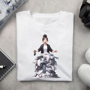 Puede incluir: Una camiseta blanca con un gr&aacute;fico de una mujer de pie frente a una gran pila de ropa. La mujer tiene el pelo oscuro y lleva una chaqueta negra. La pila de ropa est&aacute; compuesta por varios colores y patrones.