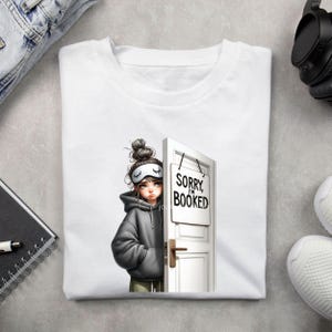 Puede incluir: Camiseta blanca con una ilustraci&oacute;n de dibujos animados de una persona con antifaz y sudadera con capucha, asom&aacute;ndose por detr&aacute;s de una puerta blanca con un letrero que dice "Sorry I'm Booked". La persona tiene el pelo recogido en un mo&ntilde;o.