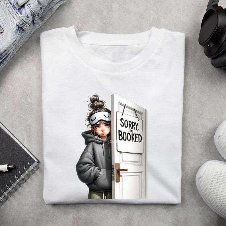 Puede incluir: Camiseta blanca con una ilustraci&oacute;n de dibujos animados de una persona con antifaz y sudadera con capucha, asom&aacute;ndose por detr&aacute;s de una puerta blanca con un letrero que dice "Sorry I'm Booked". La persona tiene el pelo recogido en un mo&ntilde;o.