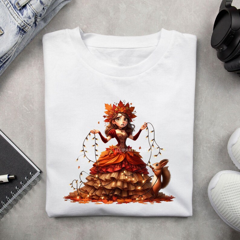 May include: White t-shirt featuring a whimsical illustration of a woman in an autumn-themed dress, adorned with a leaf crown and holding string lights. A squirrel is next to her. The design has warm colors of orange, brown, and gold.