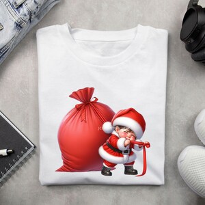 May include: White t-shirt featuring a cartoon Santa Claus character pulling a red ribbon. A large red sack is behind the Santa Claus character. The Santa Claus character is wearing a red and white suit and hat. The shirt is on a light gray surface.