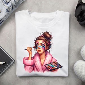 K&ouml;nnte beinhalten: Ein wei&szlig;es T-Shirt mit einer bunten Illustration einer Frau, die sich schminkt. Die Frau hat braunes Haar in einem Dutt und tr&auml;gt einen rosa Morgenmantel. Sie h&auml;lt einen Make-up-Pinsel und eine bunte Lidschattenpalette.