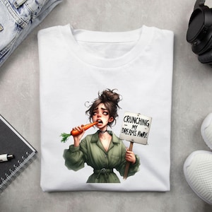 May include: White t-shirt with a cartoon illustration of a woman eating a carrot and holding a sign that says "Crunching my dreams away."
