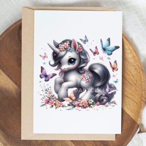 May include: A greeting card featuring a whimsical illustration of a unicorn with a silver mane and tail, adorned with a floral crown and surrounded by colorful butterflies. The unicorn has large blue eyes and is set against a white background.