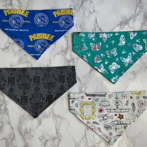 May include: Four different dog bandanas. One is blue with the words "Pawnee Non-Essential Employee" and a seal. One is green with a butterfly pattern. One is gray with a black pattern. One is white with a colorful pattern of a coffee cup, a fountain, a car, and a picture frame.