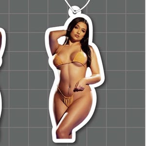 May include: Two identical air fresheners featuring a photographic print of a person with long dark hair wearing a mustard yellow bikini. Each air freshener has a white hanging string and measures 95 mm in height and 41.511 mm in width.