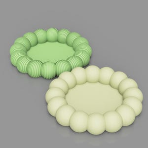Op de afbeelding: Twee decoratieve, cirkelvormige objecten. Eén is lichtgroen met een geribbelde, bolvormige rand. De andere heeft een vergelijkbaar ontwerp, maar in een lichtgeelgroene kleur met een gladde, bolvormige rand. Beide hebben een plat, cirkelvormig midden.