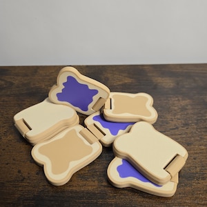 May include: A collection of wooden toy bread slices, some with tan bread and purple filling, others with tan bread and no filling. The pieces are scattered on a dark brown wooden surface.