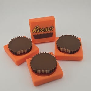 May include: Four orange square-shaped objects with a dark brown, circular top, resembling a peanut butter cup. One square has the text "Reese's" in yellow. The objects are arranged on a white surface.