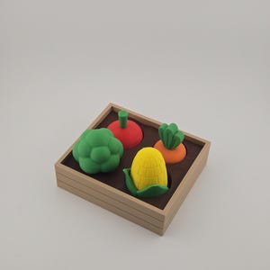 May include: A wooden tray filled with colorful toy vegetables. The set includes a green broccoli, a red tomato, an orange carrot, and a yellow corn on the cob. The vegetables are made of a soft material.