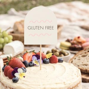 May include: A gluten-free cake topped with fresh strawberries, blueberries, raspberries, and edible flowers. A sign with the words "GLUTEN FREE" is placed on top of the cake. The cake is part of a picnic spread.