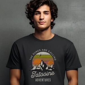 May include: A black t-shirt with a vintage-style graphic of mountains and a sunset. The text "The Dunes Are Calling Tatooine Adventures" is printed on the shirt.
