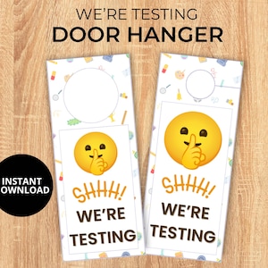 Può includere: Un appendiporta bianco con un'emoji gialla con un dito sulla bocca. Il testo "SHHH! WE'RE TESTING" è stampato in nero sull'appendiporta.