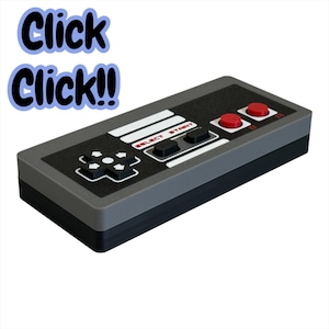 May include: A gray and black rectangular controller with red buttons and a directional pad. The text "Click Click!!" is in a blue bubble in the upper left corner. The controller has the words "SELECT START" printed on it.