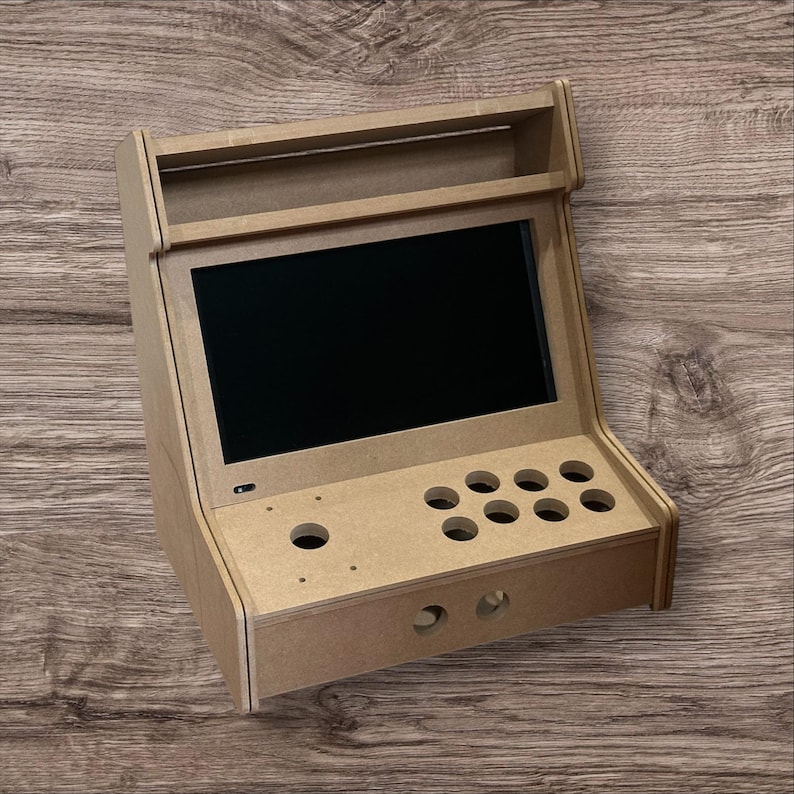 May include: A small, unfinished wooden arcade cabinet with a black screen, eight button holes, and two joystick holes. The cabinet has a rectangular shape with a top shelf and is designed for a retro gaming experience.