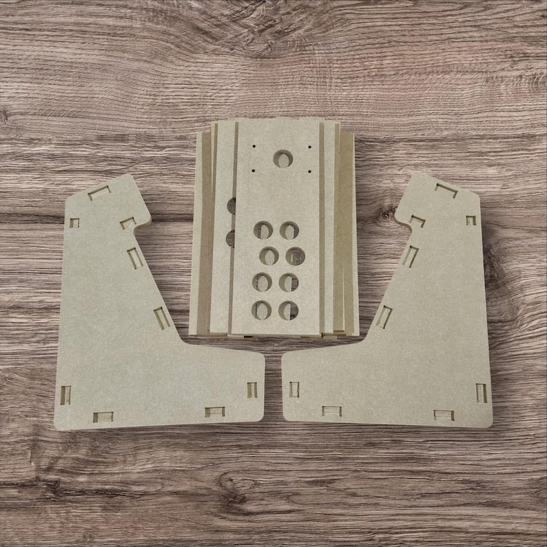 May include: Unassembled light brown wooden arcade game kit. The kit includes side panels, a control panel, and other components with pre-cut holes and slots for assembly. The pieces are arranged on a wooden surface.
