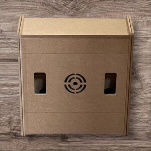 May include: A light brown, rectangular wooden box with a speaker grill and two rectangular openings. The box has a simple, modern design and is likely for electronics or storage.
