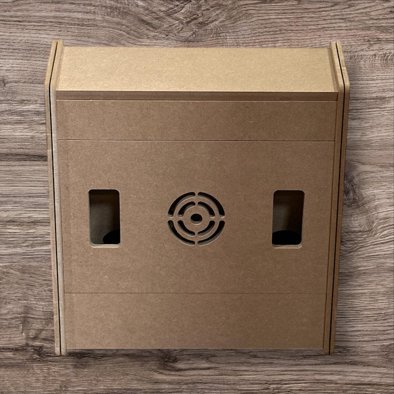 May include: A light brown, rectangular wooden box with a speaker grill and two rectangular openings. The box has a simple, modern design and is likely for electronics or storage.