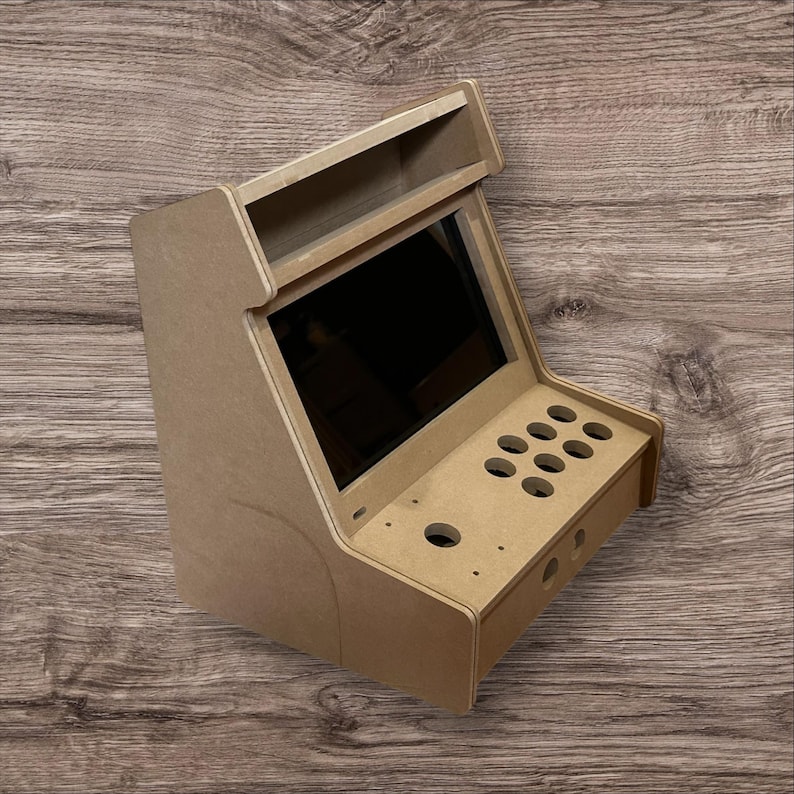 May include: A small, unfinished arcade cabinet made of light brown wood. It features a screen, button holes, and a top shelf. The cabinet is designed to house arcade game components.