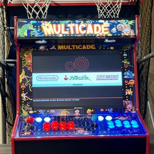 May include: A red and black Multicade arcade machine with a screen displaying a game menu. The machine has a control panel with joysticks and buttons. Above the arcade machine are two basketball hoops.