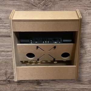 May include: A light brown, unfinished wooden arcade cabinet with a rectangular shape. It features a top section, a middle section with two circular speaker holes, and a bottom section. The cabinet houses a black electronic component.