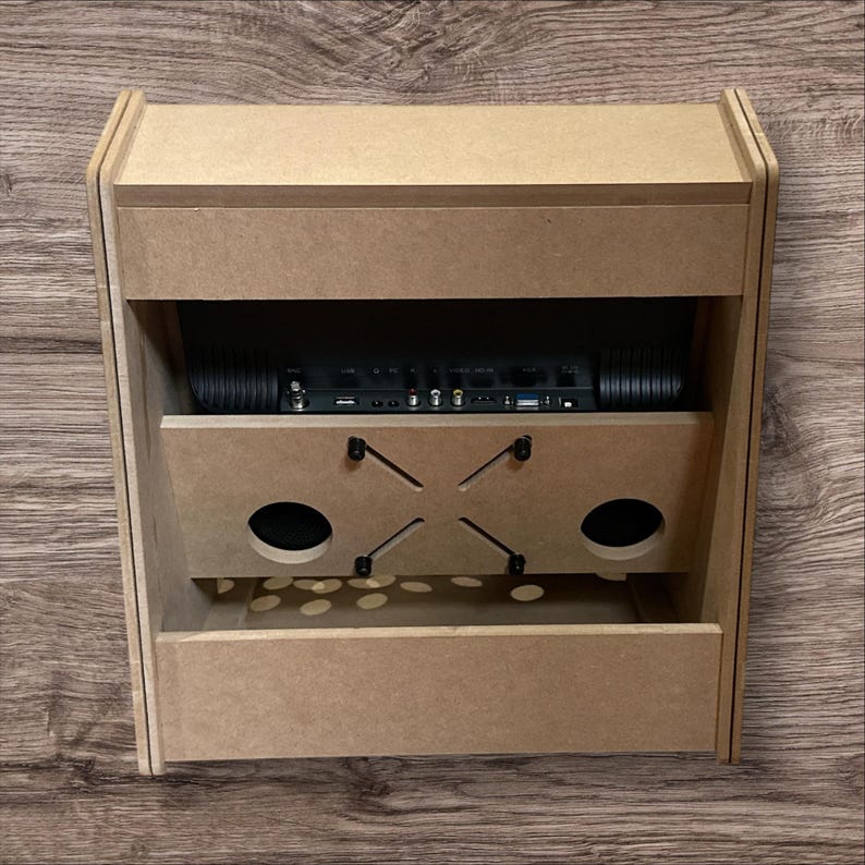 May include: A light brown, unfinished wooden arcade cabinet with a rectangular shape. It features a top section, a middle section with two circular speaker holes, and a bottom section. The cabinet houses a black electronic component.