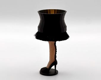 Pode incluir: Uma lâmpada de novidade com uma abajur preto e uma franja na parte inferior. A lâmpada é suportada por uma perna usando uma meia de rede e um sapato de salto alto preto. A base da lâmpada é preta e redonda.
