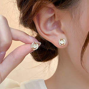 Könnte beinhalten: Goldfarbene Ohrstecker mit einem zentralen, perlmuttweißen Stein, umgeben von einem Kranz aus kleinen, klaren Kristallen. Die Ohrringe sind rund mit einem doppelten Goldrahmen. Ein Ohrring wird ans Ohr gehalten, der andere ist bereits eingesetzt.