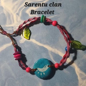 May include: A handmade bracelet with the text "Sarentu clan Bracelet". The bracelet features a twisted red and brown cord, adorned with pink beads, green leaf charms, and a turquoise circular bead with a white design.
