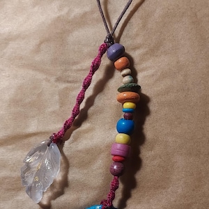 May include: A handmade pendant featuring a clear leaf-shaped charm, a strand of colorful wooden beads, and a blue, round painted charm. The pendant is strung on a brown cord with a twisted red accent.